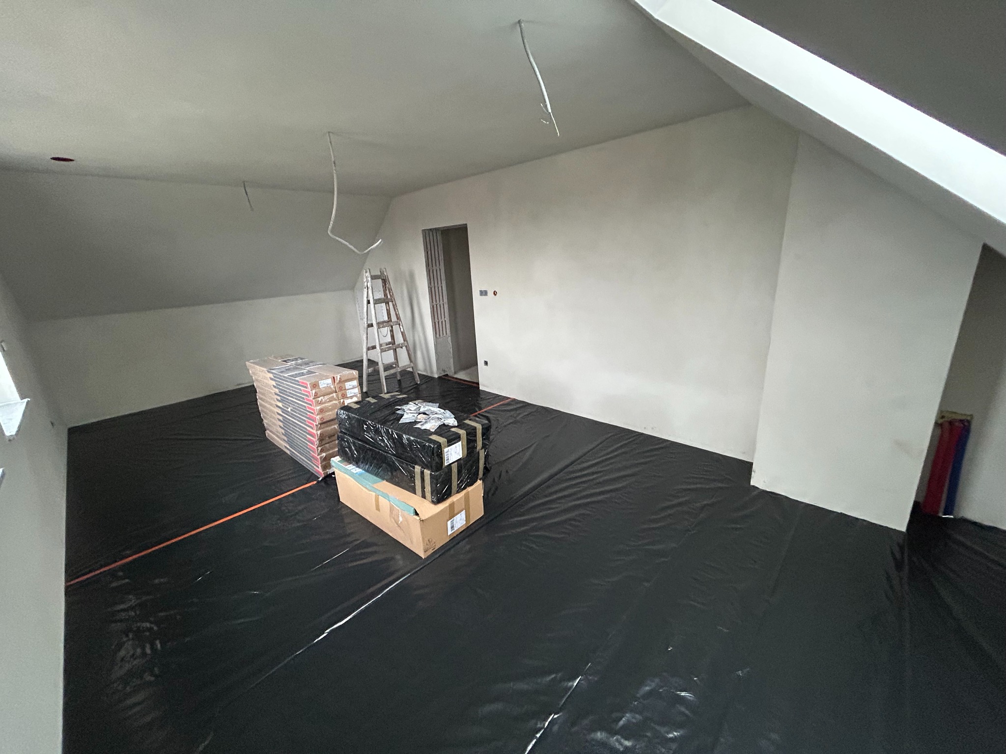 Poddasze w trakcie przygotowania do prac wykończeniowych: zabezpieczona podłoga folią, materiały budowlane, drabina. Widoczne kable wystające z sufitu.
