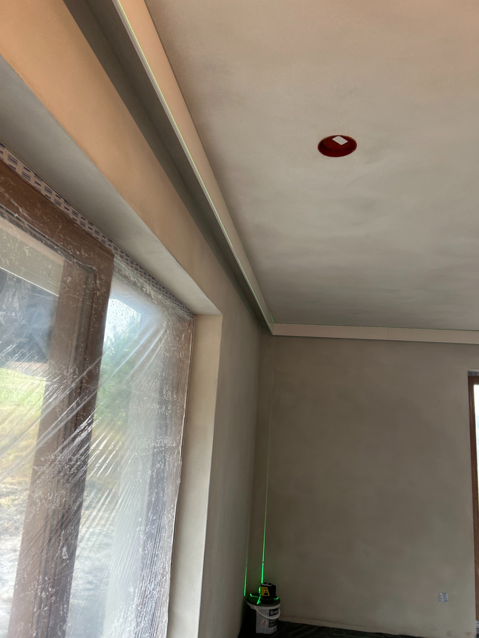 Wygładzone ściany i sufit w pokoju, okno zabezpieczone folią, widoczny laser krzyżowy. Przygotowanie do malowania.