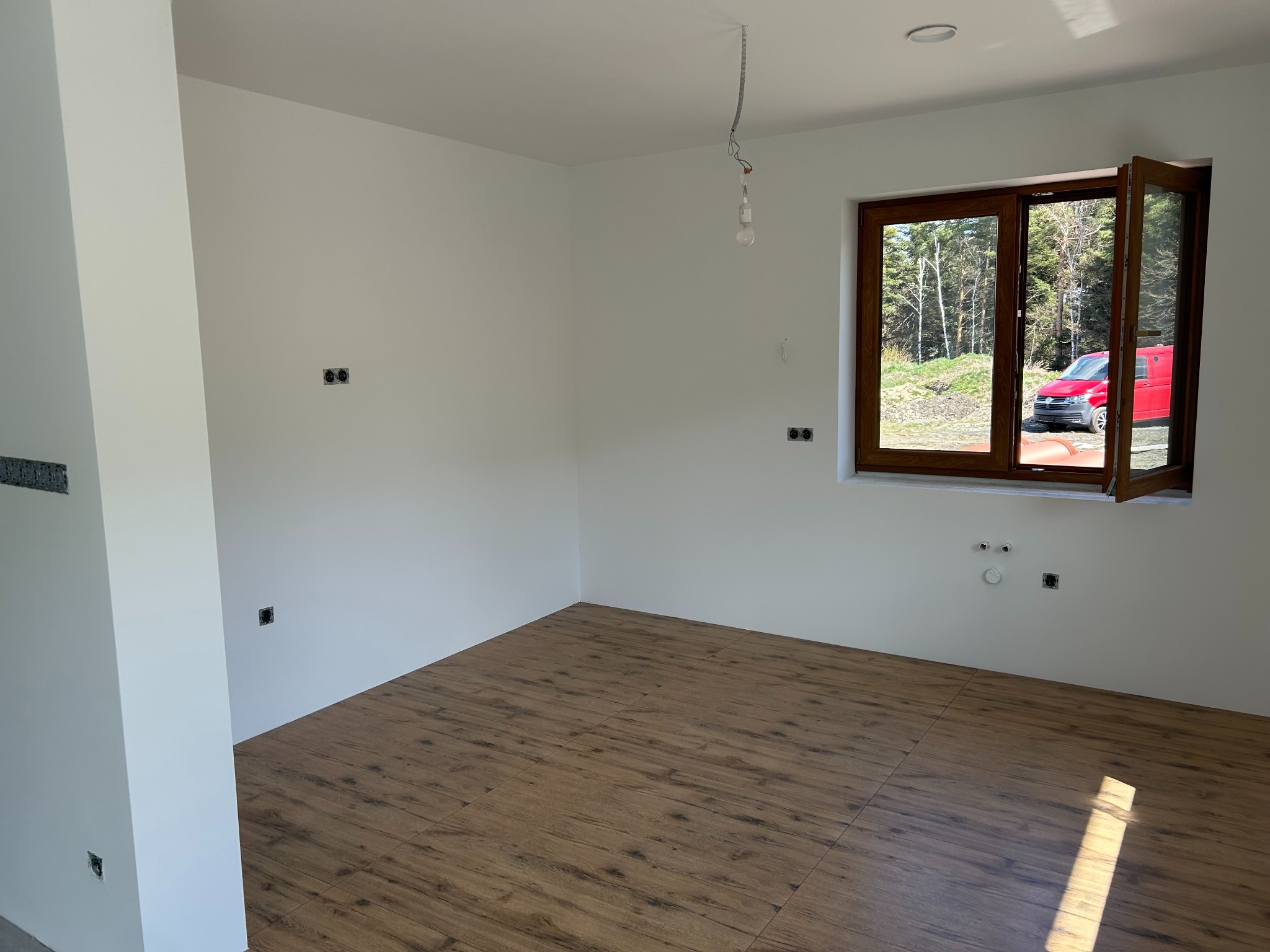 Pusty pokój po remoncie: białe ściany, drewnopodobna podłoga, otwarte okno z widokiem na zieleń. Widoczne gniazdka elektryczne i wystające kable oświetleniowe.