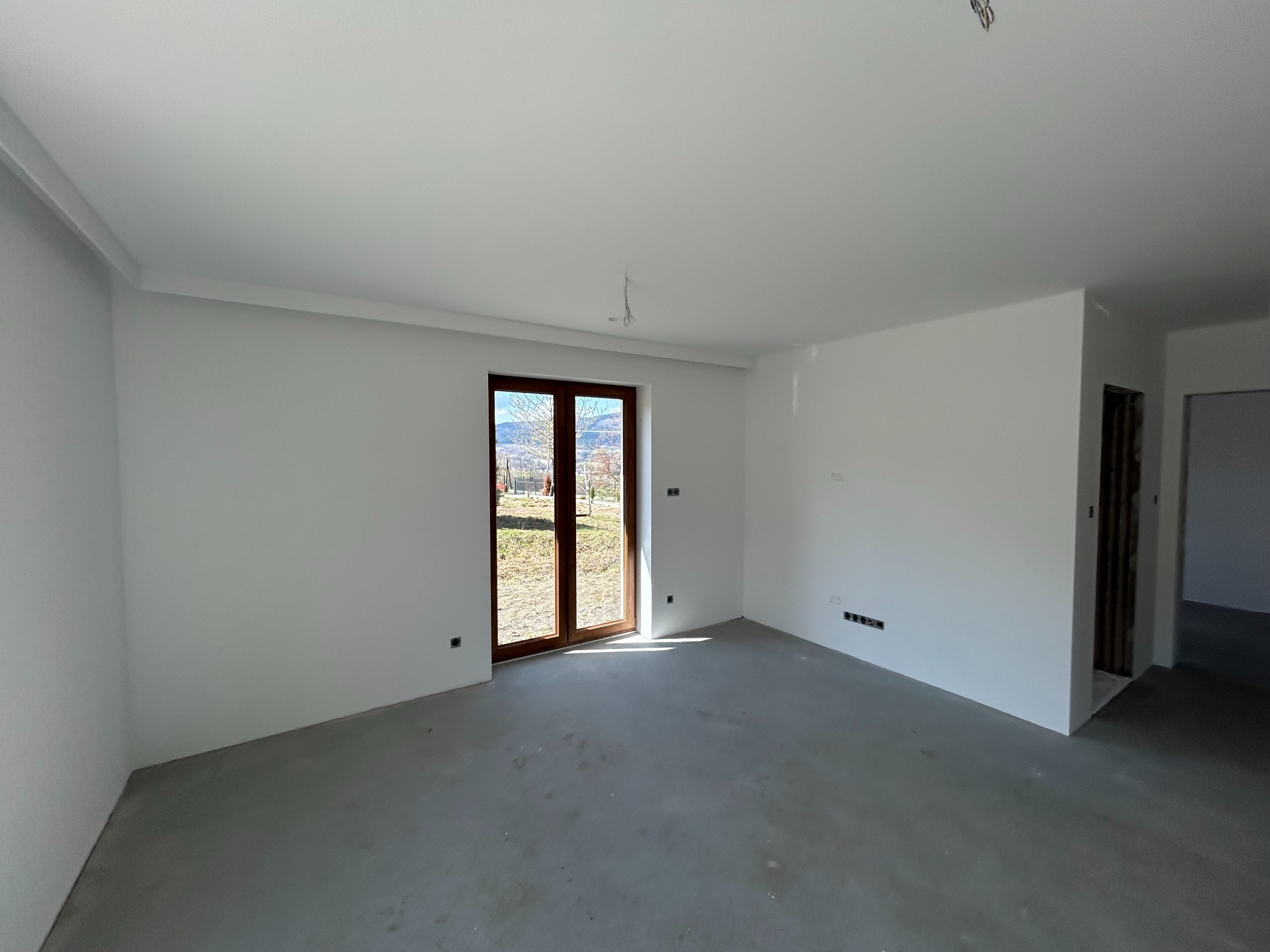 Pusty, jasny pokój z gładkimi, białymi ścianami i szarą podłogą. Widoczne drewniane drzwi balkonowe z widokiem na zewnątrz. Przygotowany do dalszych prac wykończeniowych.