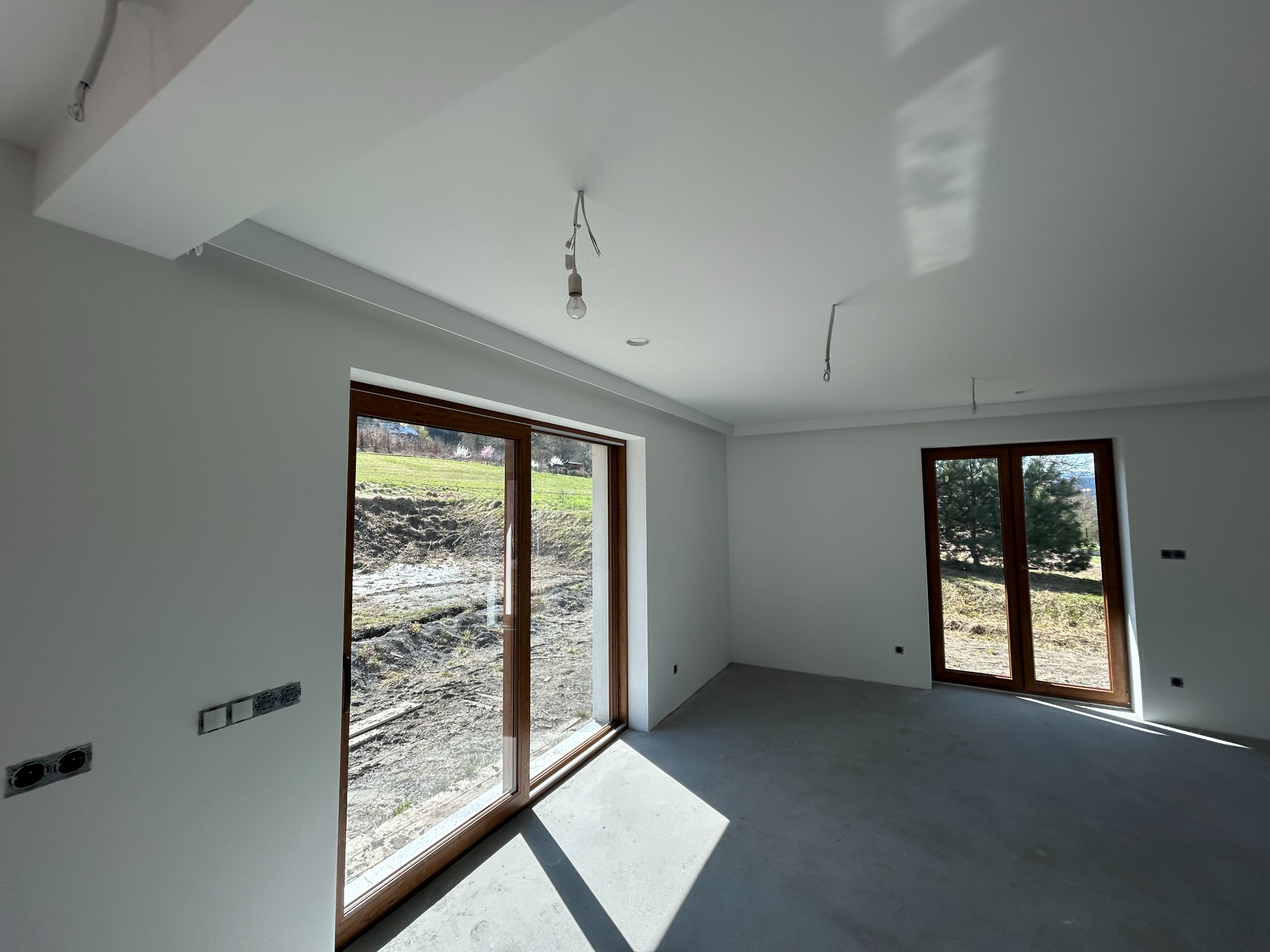 Jasne, świeżo pomalowane wnętrze z dużymi oknami i surową podłogą. Widoczne gniazdka elektryczne i wiszące kable, sugerujące etap wykończeniowy.