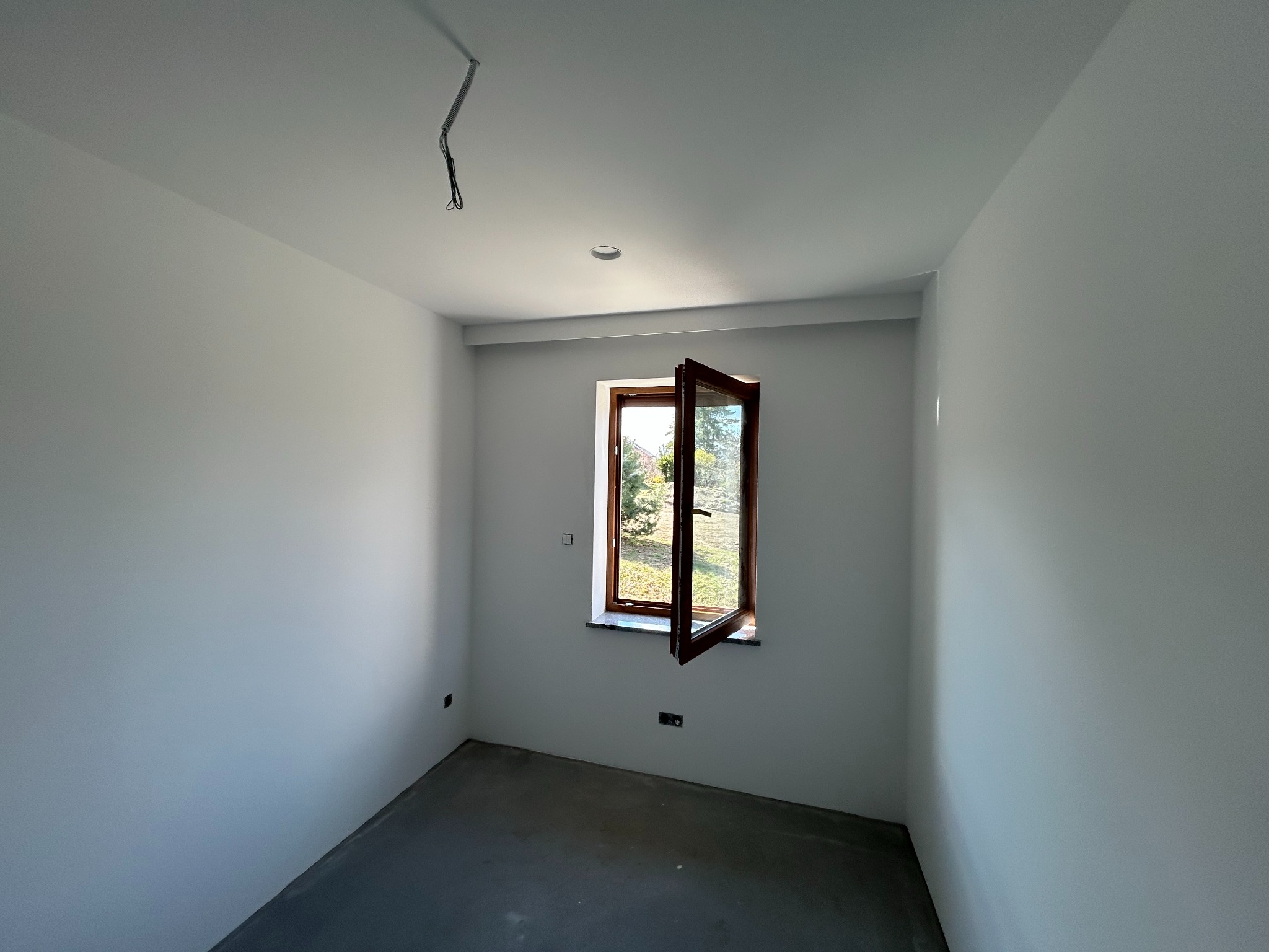 Pomalowane na biało wnętrze pokoju z otwartym oknem, przez które widać zieleń. Na suficie widoczne przewody i zamontowane oświetlenie. Szara podłoga.