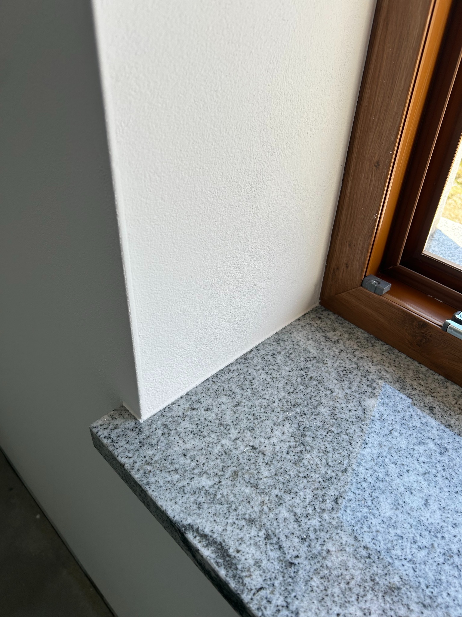 Gładka, biała ściana z oknem o drewnianej ramie i szarym, granitowym parapetem. Widoczny fragment podłogi. Minimalistyczne wykończenie wnętrza.