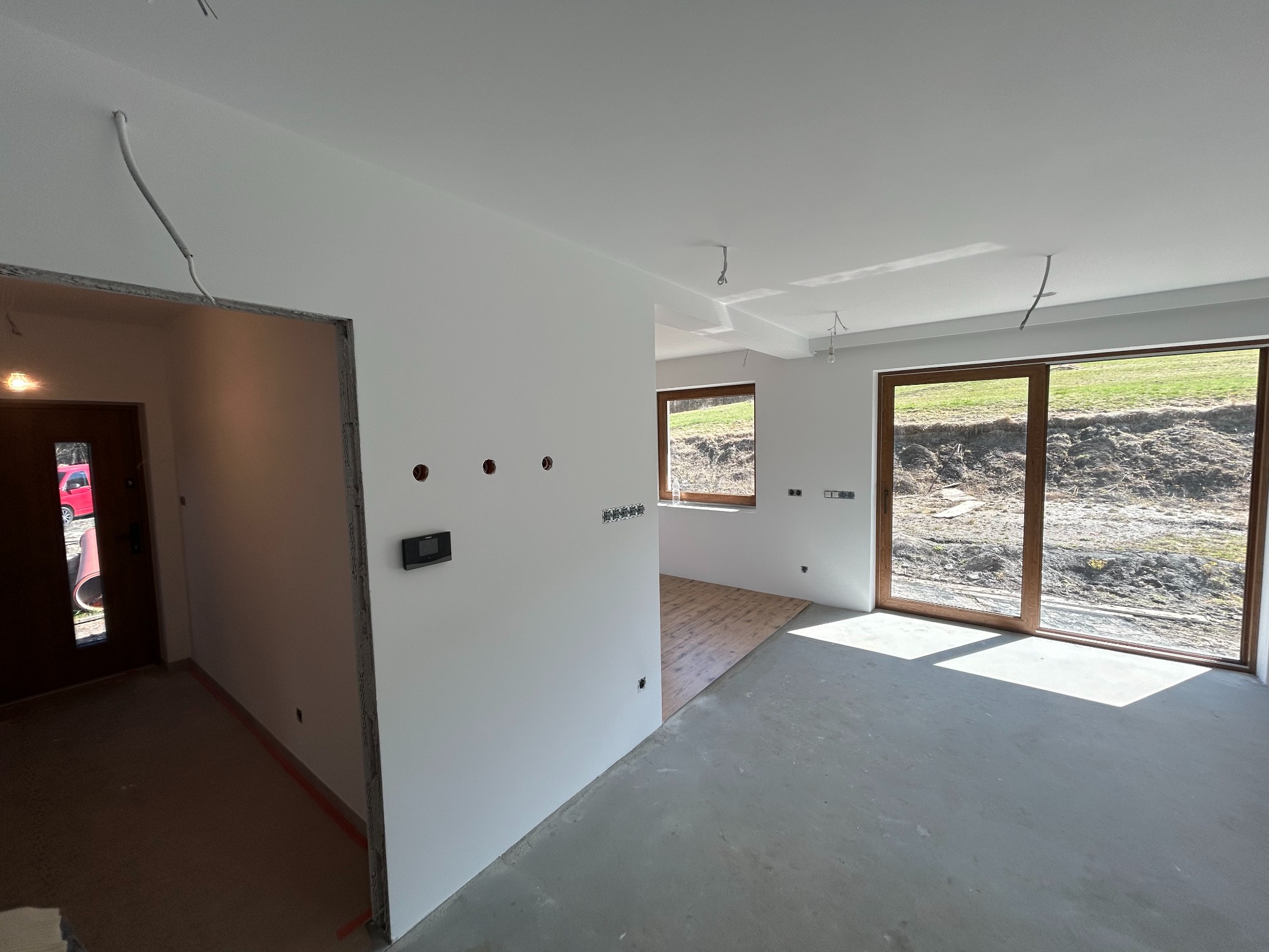 Wnętrze w stanie deweloperskim z białymi ścianami, surową podłogą i częściowo ułożonym parkietem. Widoczne instalacje elektryczne i okna z widokiem na zewnątrz.