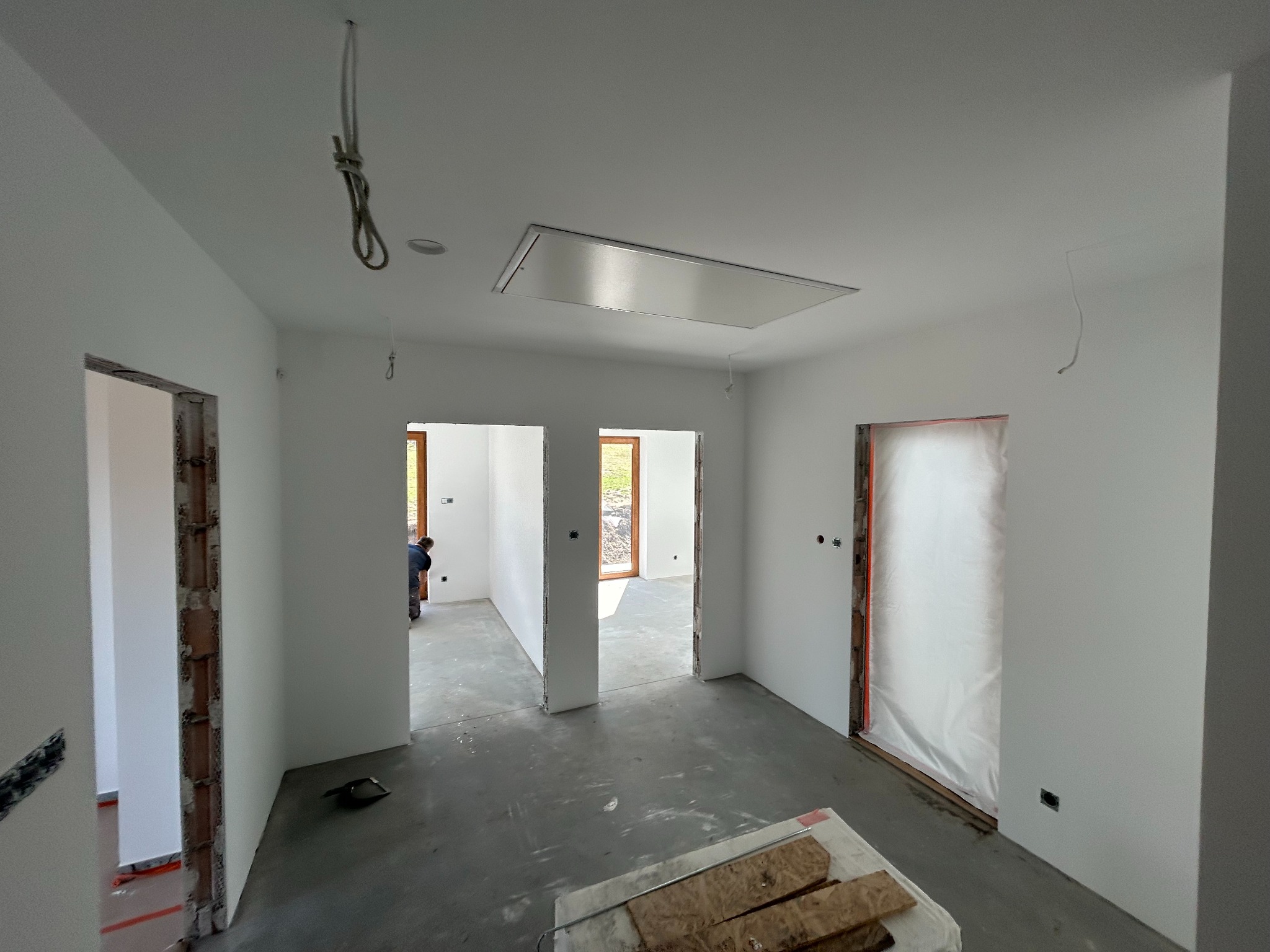 Wnętrze w trakcie malowania na biało, widoczne przygotowane otwory drzwiowe, surowa podłoga i wystające kable. Prace wykończeniowe w nowym domu.
