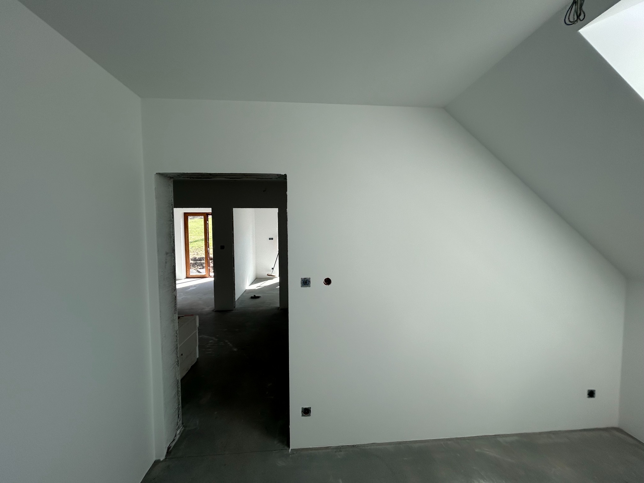 Białe, świeżo pomalowane poddasze ze skosami i widocznymi gniazdkami elektrycznymi. W tle widoczne kolejne pomieszczenie przez otwarte drzwi.