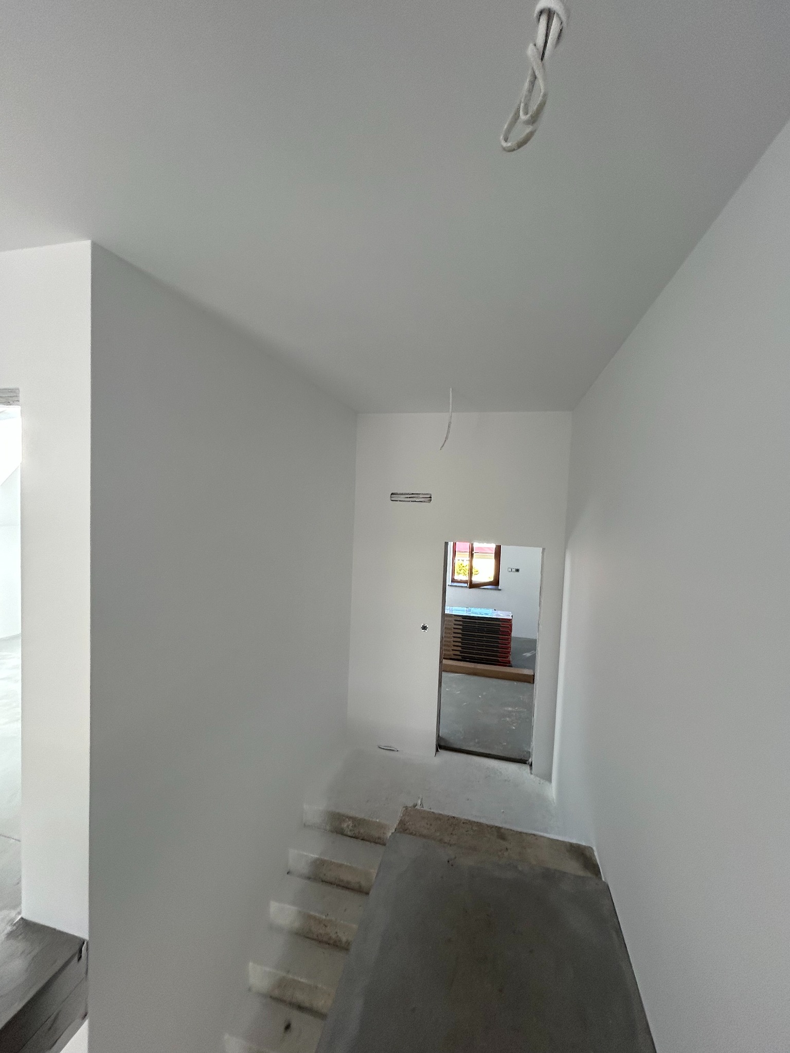 Białe, świeżo pomalowane ściany klatki schodowej w domu jednorodzinnym. Widoczne schody w surowym stanie i fragment okna w tle. Minimalistyczny efekt po malowaniu.