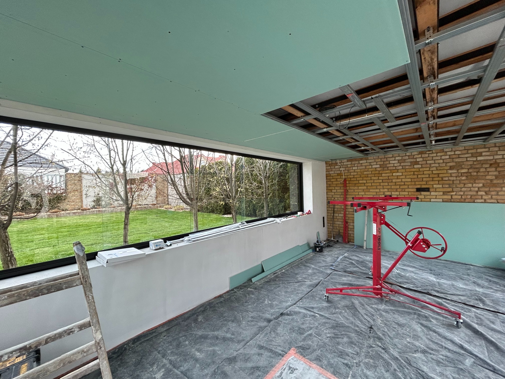 Montaż płyt gipsowo-kartonowych na suficie w pomieszczeniu z dużym oknem. Widoczny podnośnik do płyt i metalowa konstrukcja sufitu podwieszanego.