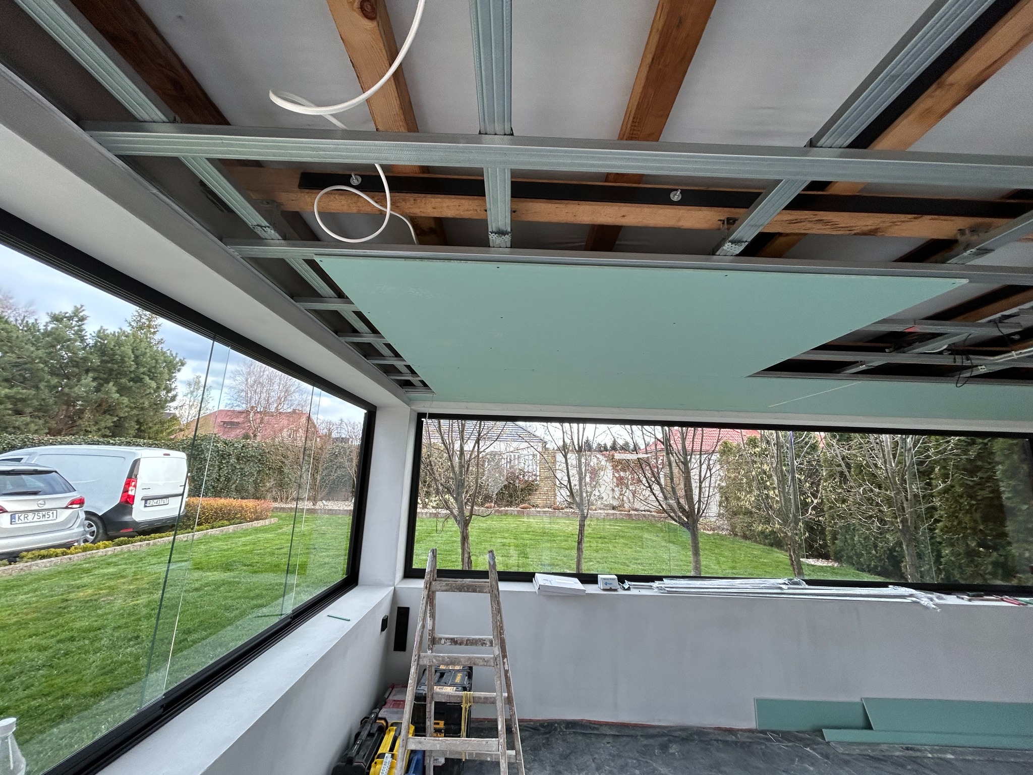 Montaż sufitu podwieszanego z płyt gipsowo-kartonowych w jasnym pomieszczeniu z dużymi oknami. Widoczna drabina i narzędzia. Etap prac wykończeniowych.