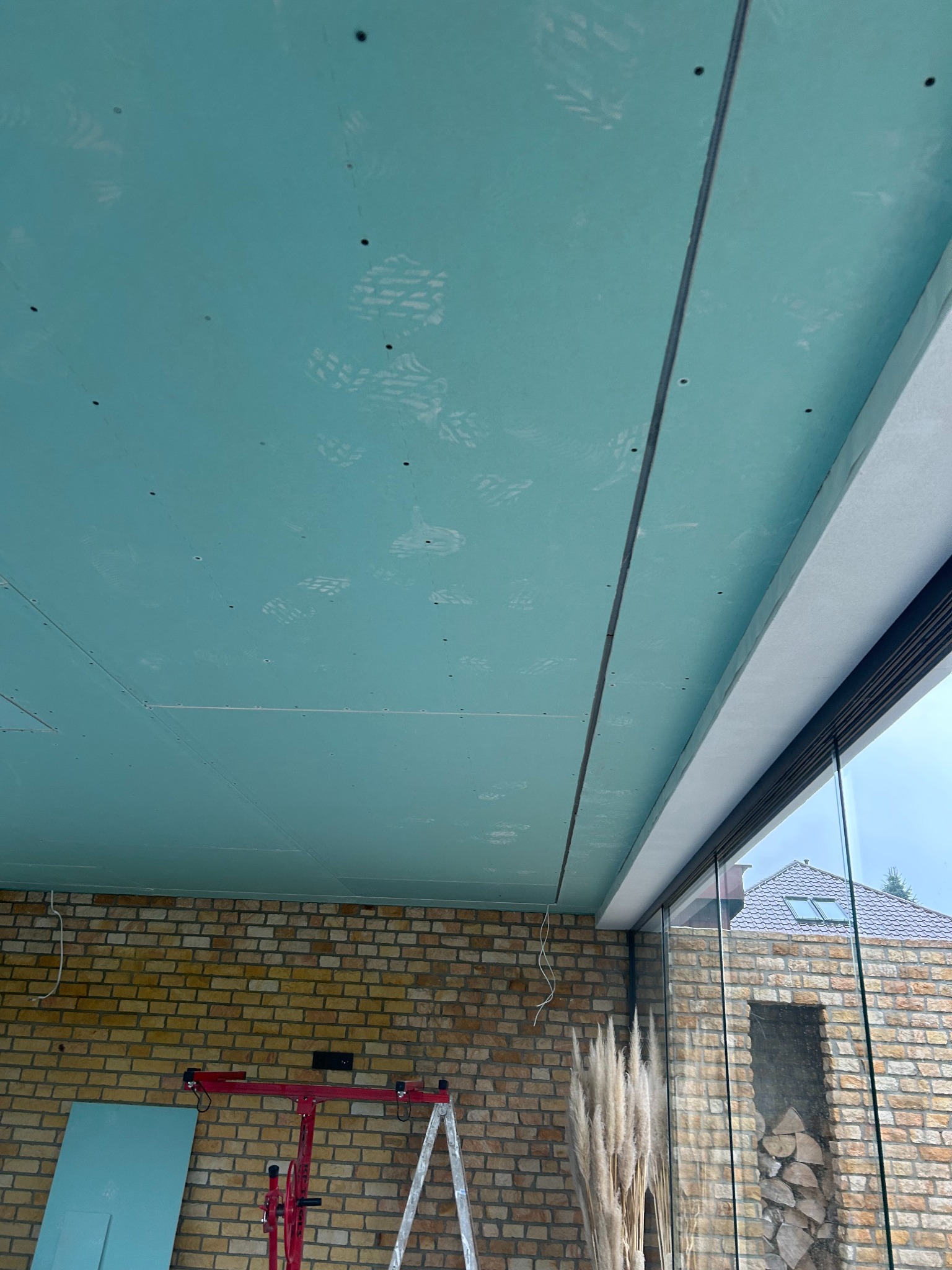 Montaż płyt gipsowo-kartonowych na suficie w pomieszczeniu z ceglaną ścianą i dużym oknem. Widoczne ślady stóp na płytach i podnośnik do płyt.