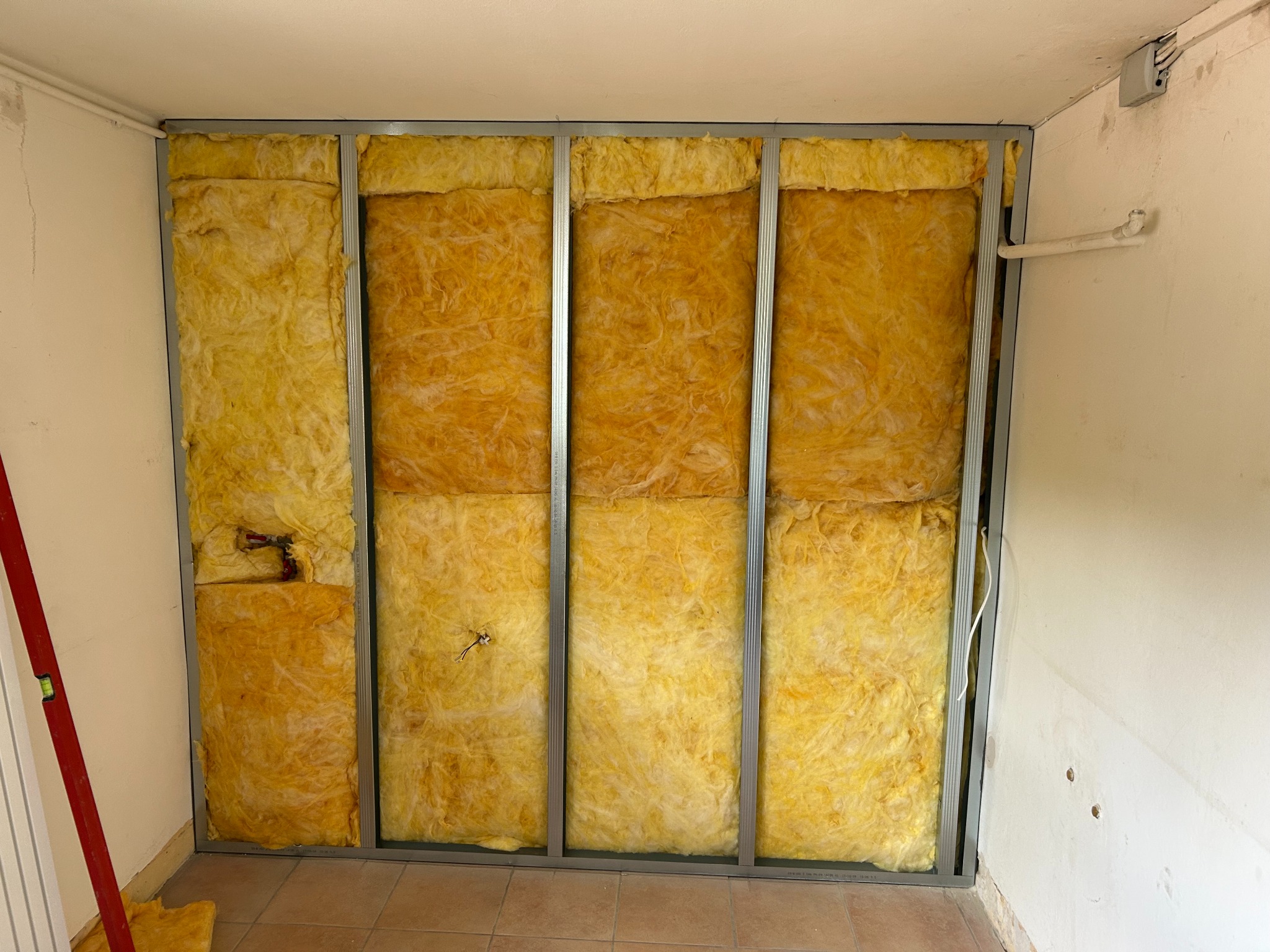 Montaż izolacji z wełny mineralnej w metalowym stelażu na ścianie. Widoczne fragmenty instalacji elektrycznej i poziomica.