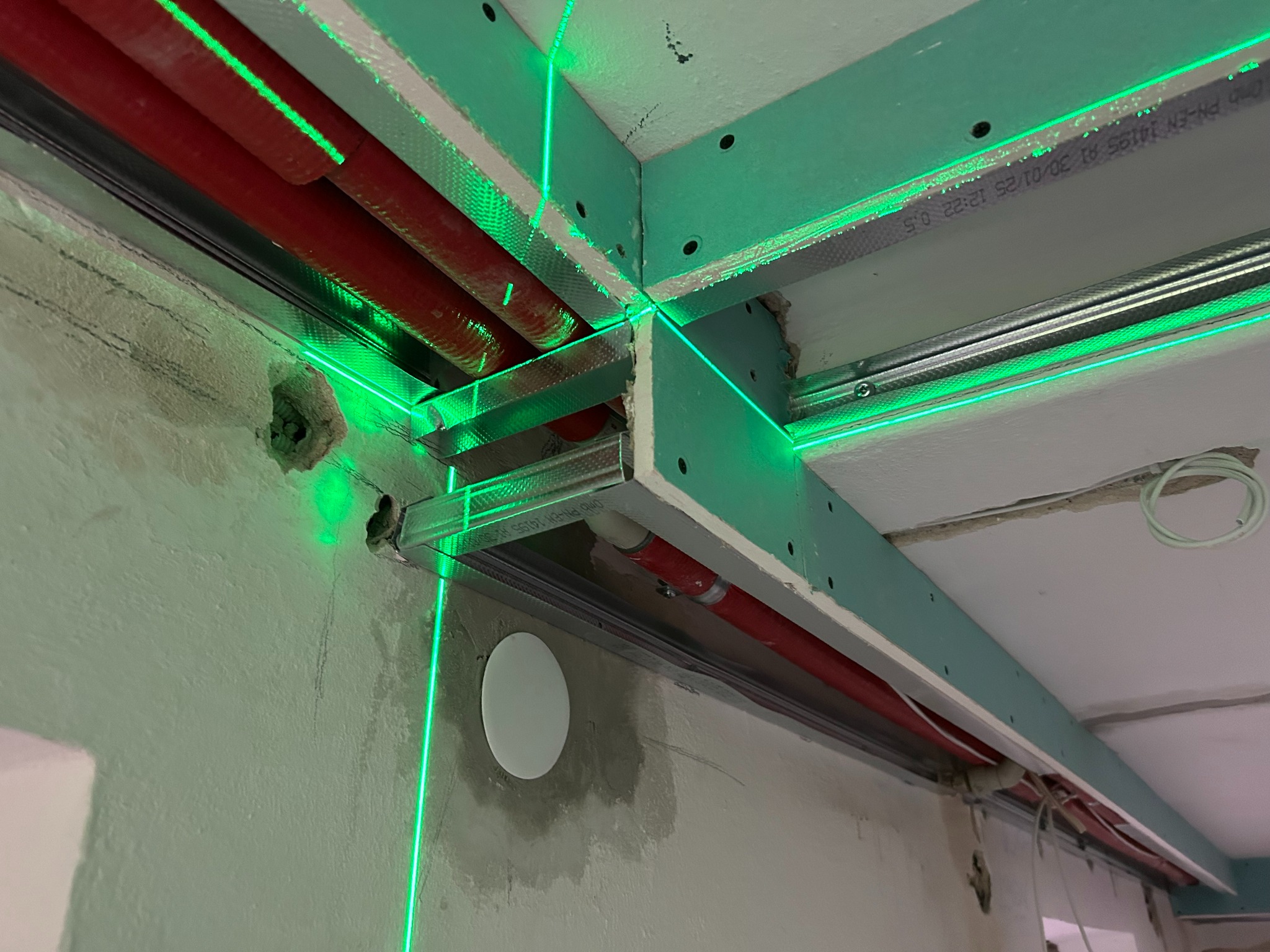 Montaż sufitu podwieszanego z płyt gipsowo-kartonowych, widoczne profile metalowe, rury i zielony laser wyznaczający poziom. Etap prac wykończeniowych.