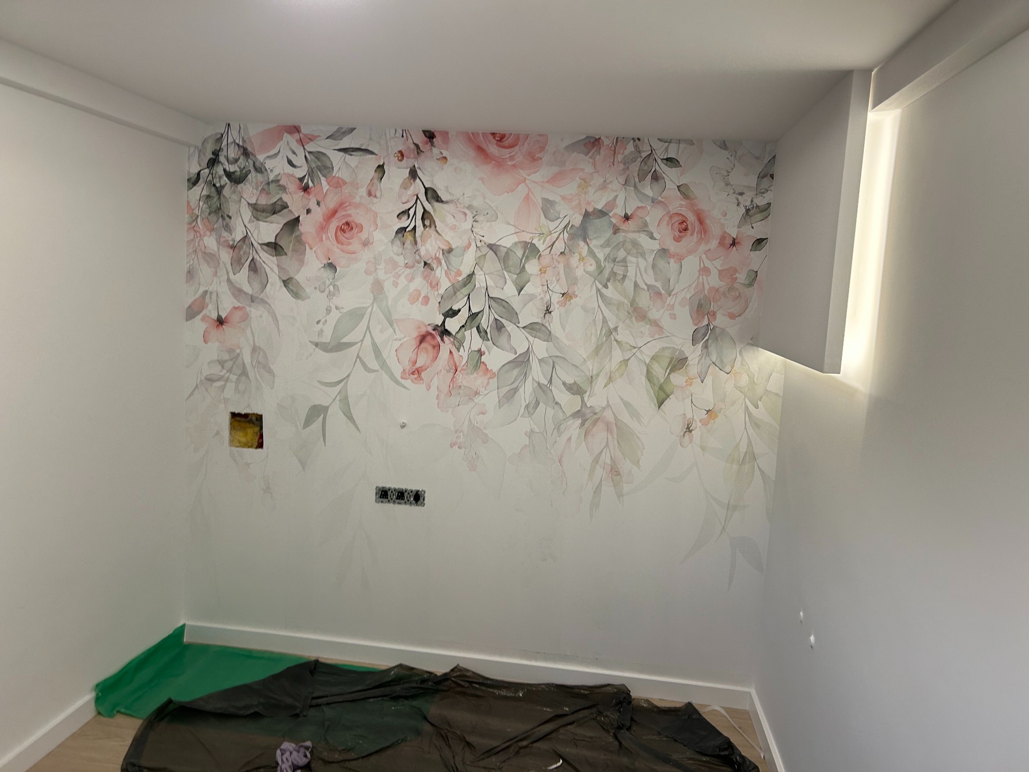 Ściana z tapetą w kwiaty, zabezpieczona folią malarską. Widoczne gniazdka elektryczne i otwór po puszce. Jasne wnętrze po remoncie, z akcentem florystycznym.