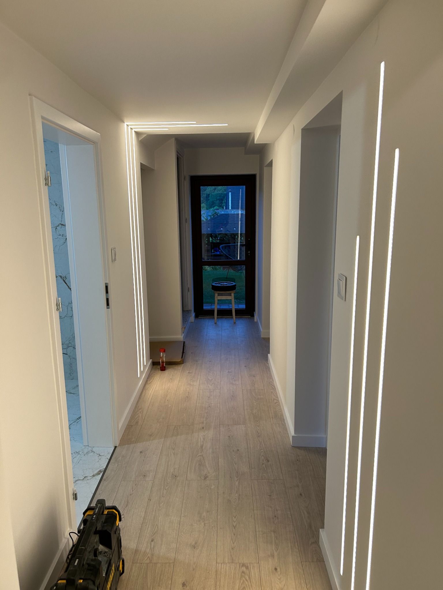 Nowoczesny korytarz z oświetleniem LED w pionowych listwach. Jasne ściany, drewnopodobna podłoga. W tle widoczne drzwi na zewnątrz i narzędzia na podłodze.