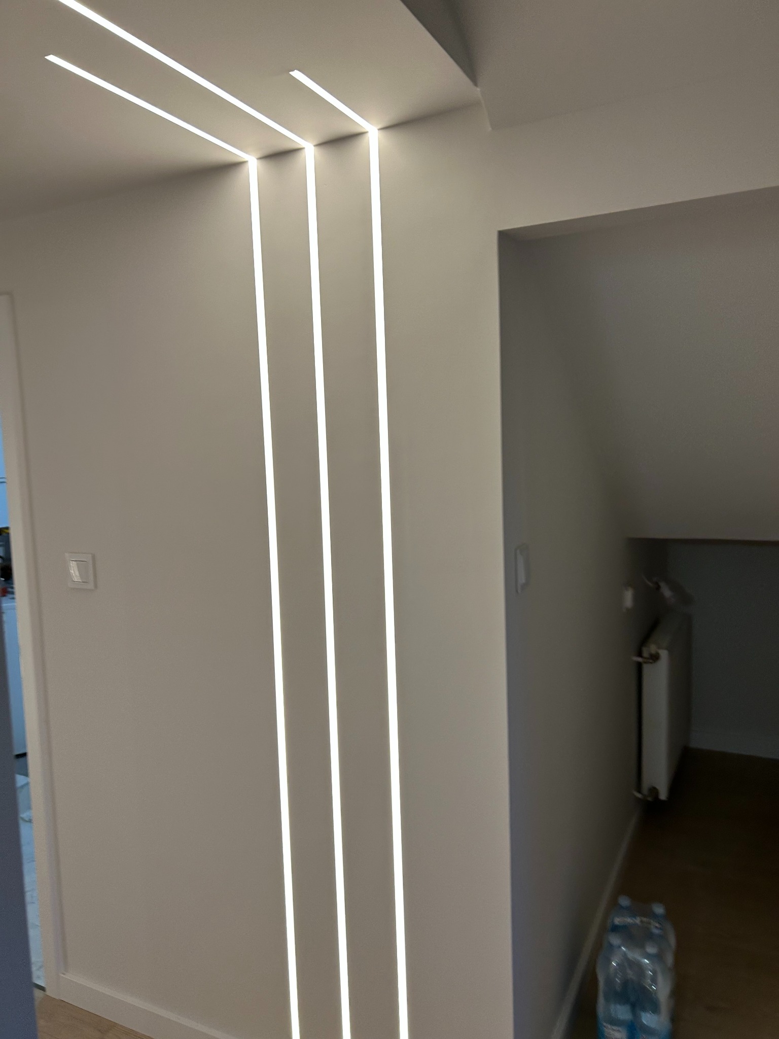 Nowoczesne oświetlenie LED wkomponowane w ścianę, tworzące pionowe linie światła. Minimalistyczny design wnętrza z białymi ścianami i drewnianą podłogą.