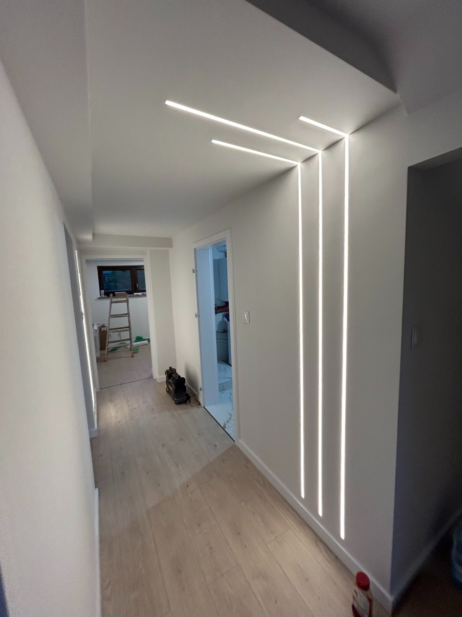 Nowoczesny korytarz z oświetleniem LED wpuszczonym w ścianę i sufit, jasne panele podłogowe, białe ściany. Widoczny fragment drabiny w tle.