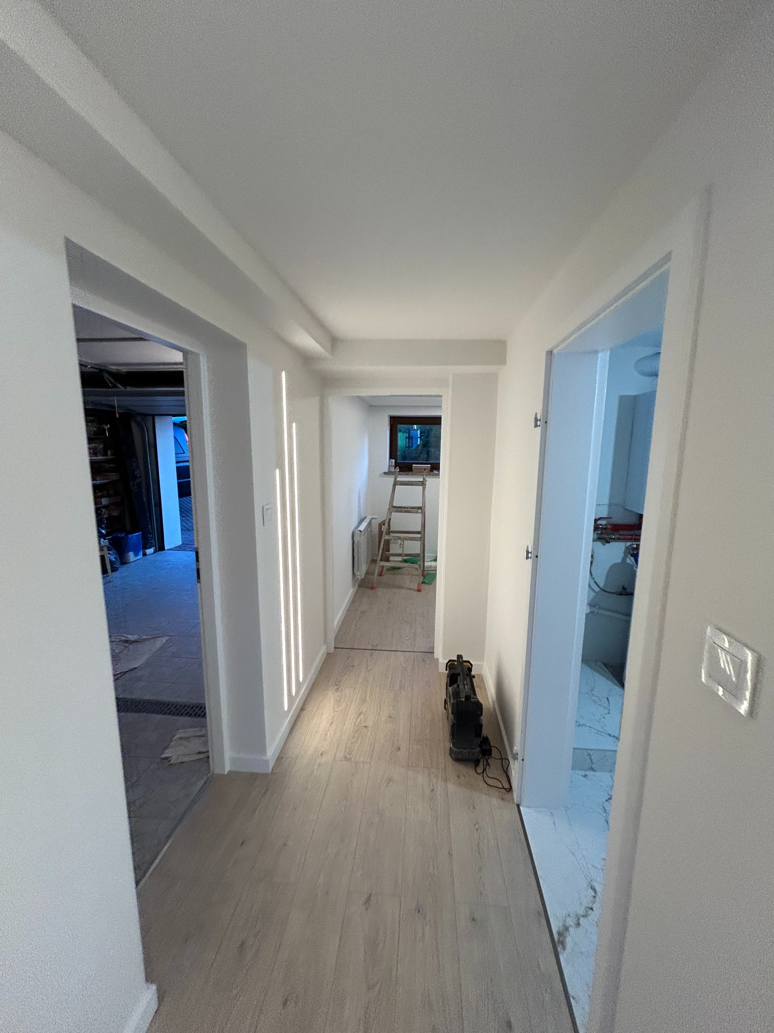 Biały korytarz po remoncie z drewnianą podłogą i oświetleniem LED w ścianach. Widoczne otwarte drzwi do garażu i łazienki z marmurową podłogą.