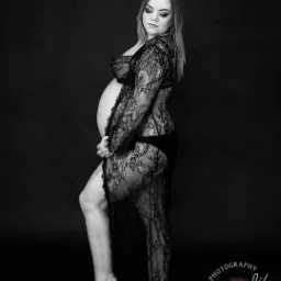 Czarno-białe zdjęcie kobiety w ciąży, pozującej bokiem w koronkowej bieliźnie i narzutce na ciemnym tle, z delikatnym uśmiechem i dłonią na brzuchu.