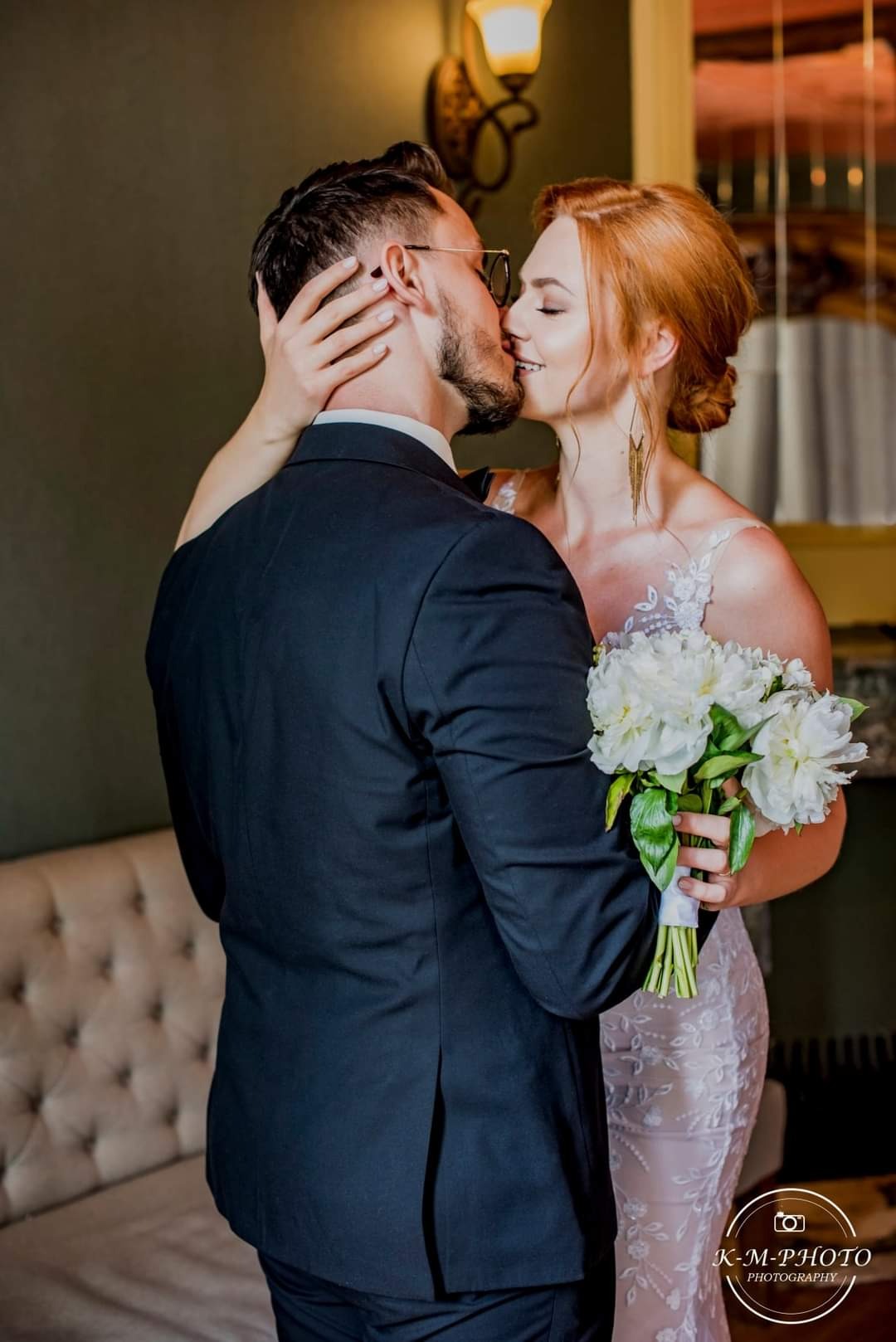 Pocałunek nowożeńców: panna młoda z rudymi włosami i bukietem białych piwonii, pan młody w okularach i ciemnym garniturze, ciepłe światło lampy w tle.