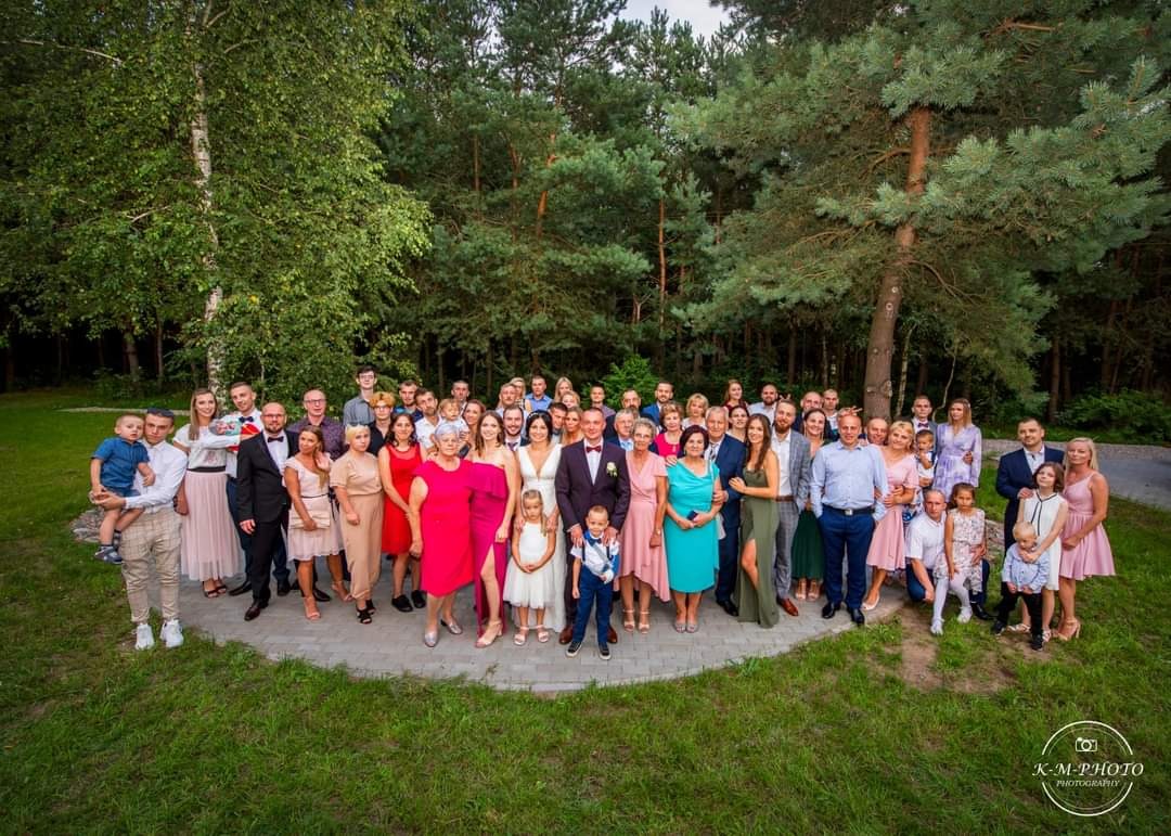 Duża grupa gości weselnych pozuje do zdjęcia grupowego na tle lasu, z parą młodą w centrum i dziećmi w pierwszym rzędzie, uchwycona z lekkiej góry.