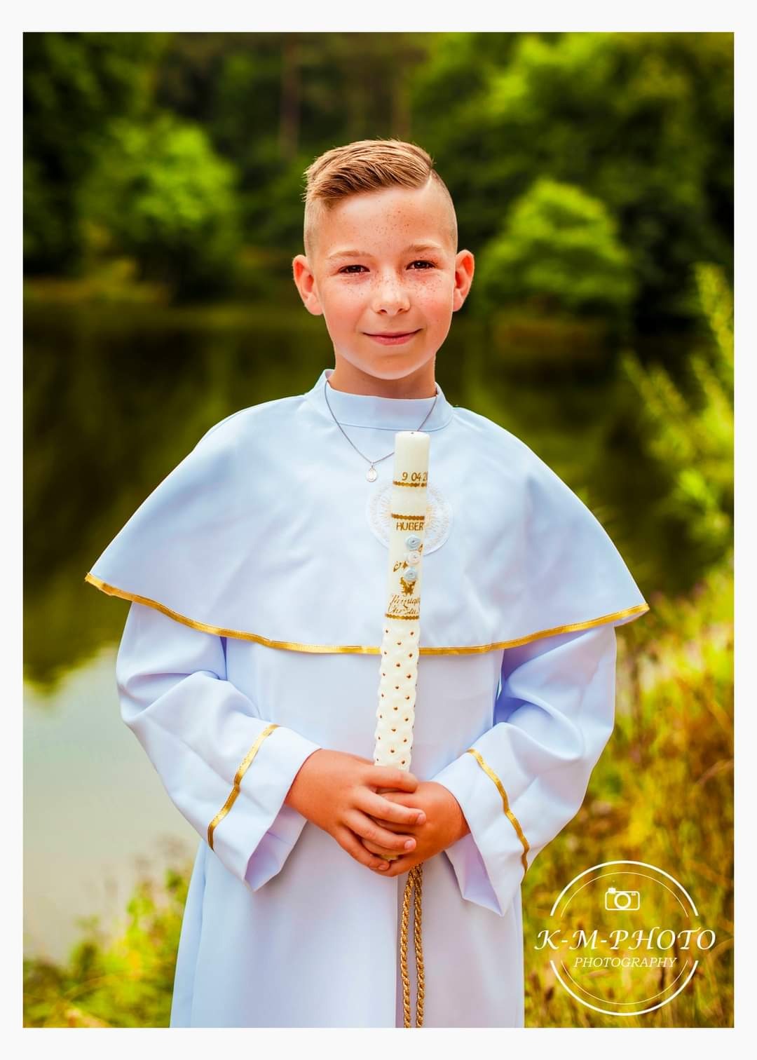 Portret chłopca w białej albie z pasem, trzymającego świecę komunijną na tle jeziora i zieleni.