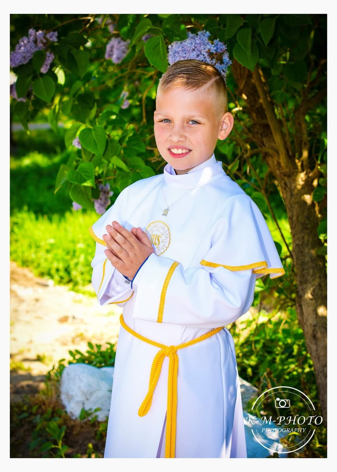 Uśmiechnięty chłopiec w białej albie z żółtym sznurem, złożone dłonie, na tle kwitnącego bzu i zieleni, z widocznym logo fotografa w prawym dolnym rogu.