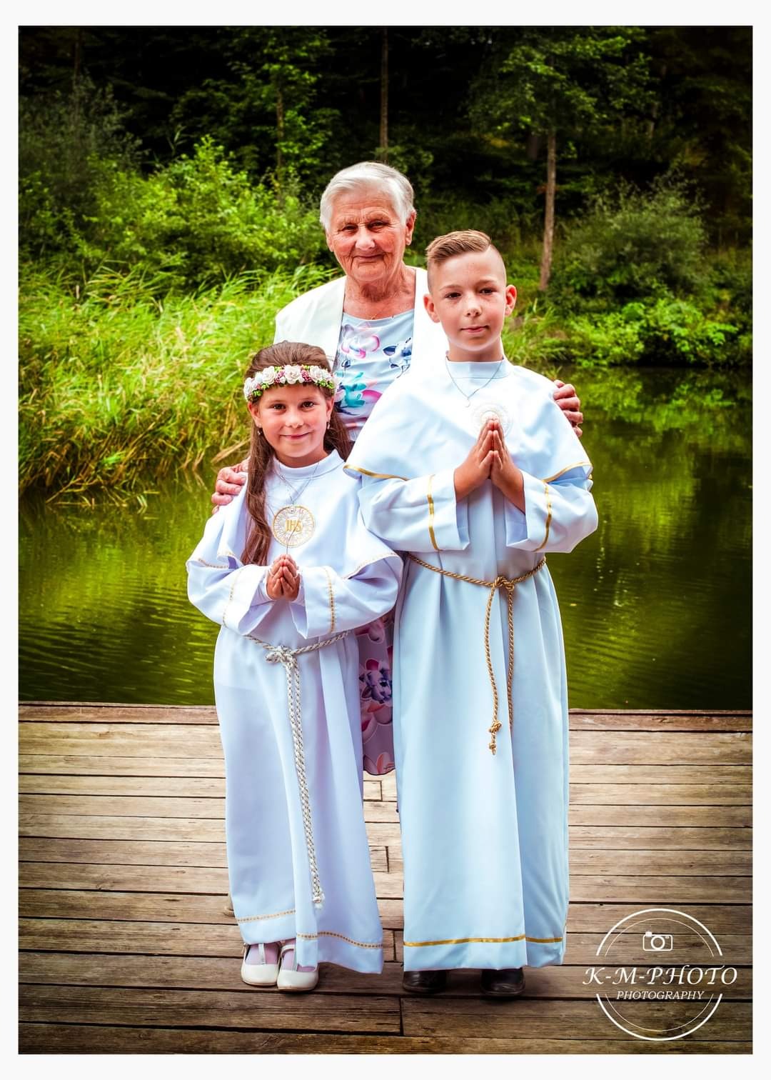 Dwoje dzieci w strojach komunijnych pozuje z babcią na drewnianym pomoście nad stawem, dziewczynka ma wianek z kwiatów na głowie.