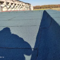 Nowy dach pokryty papą termozgrzewalną na budynku z rusztowaniami w tle, cień osoby fotografującej widoczny na powierzchni.