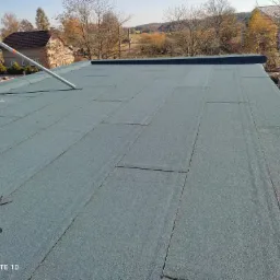 Nowo położony, płaski dach pokryty papą w kolorze szaro-zielonym, widoczny z góry, z elementami rusztowania i krajobrazem w tle.