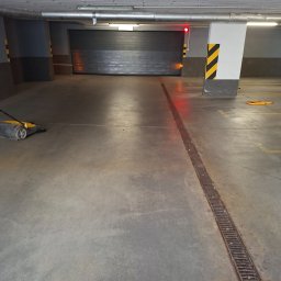 ROBSON Robert Panasiewicz - Wnętrze parkingu podziemnego z betonową posadzką i maszyną do czyszczenia podłóg. Widoczna krata odpływowa i brama garażowa z sygnalizacją świetlną.