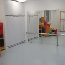 Wnętrze sali gimnastycznej z lustrami, białymi szafkami w zabudowie i kolorowymi przyborami do ćwiczeń widocznymi w odbiciu.