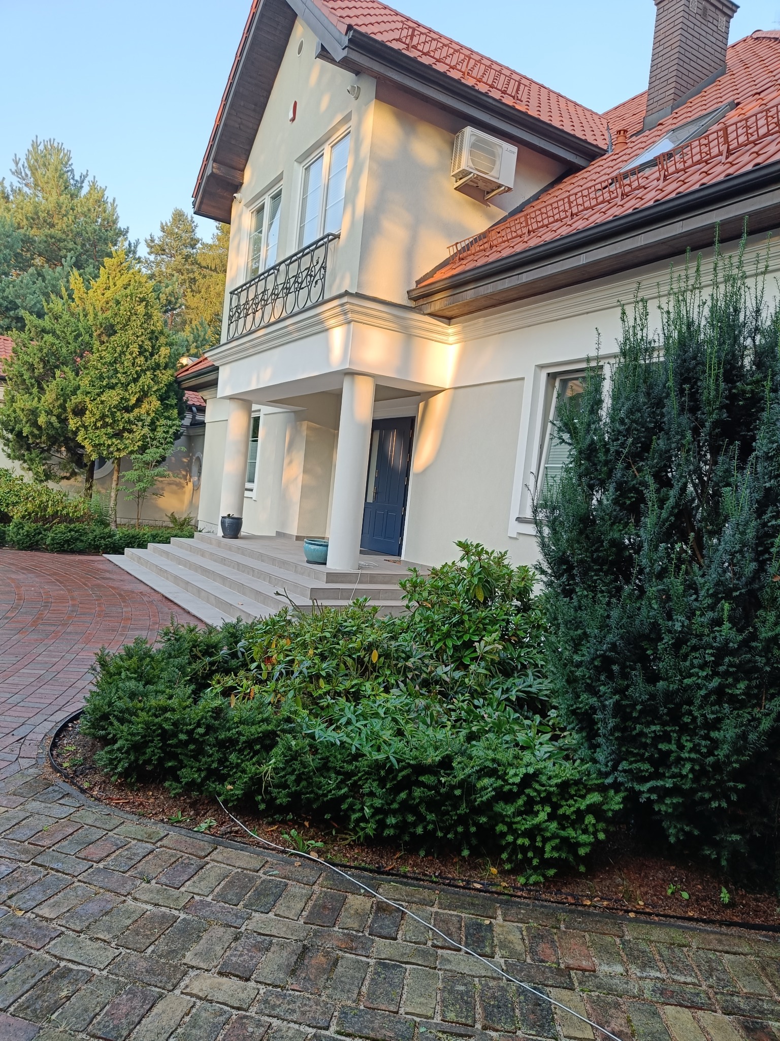 Elegancki dom jednorodzinny z kolumnami przy wejściu, zadbanym ogrodem i brukowanym podjazdem w promieniach popołudniowego słońca.