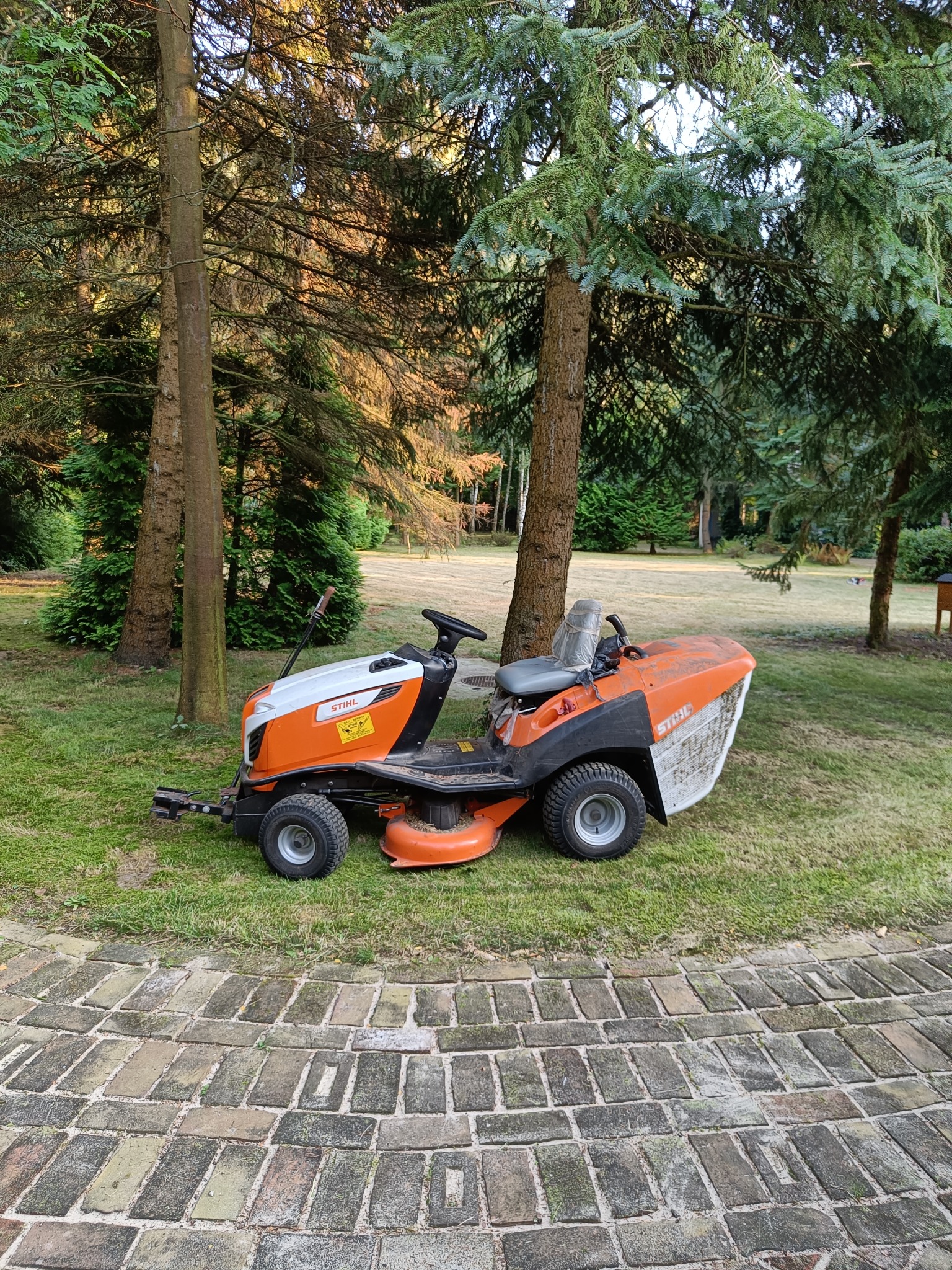 Pomarańczowo-biała kosiarka traktorek marki Stihl zaparkowana na trawniku w ogrodzie, widoczna z perspektywy brukowanej ścieżki.