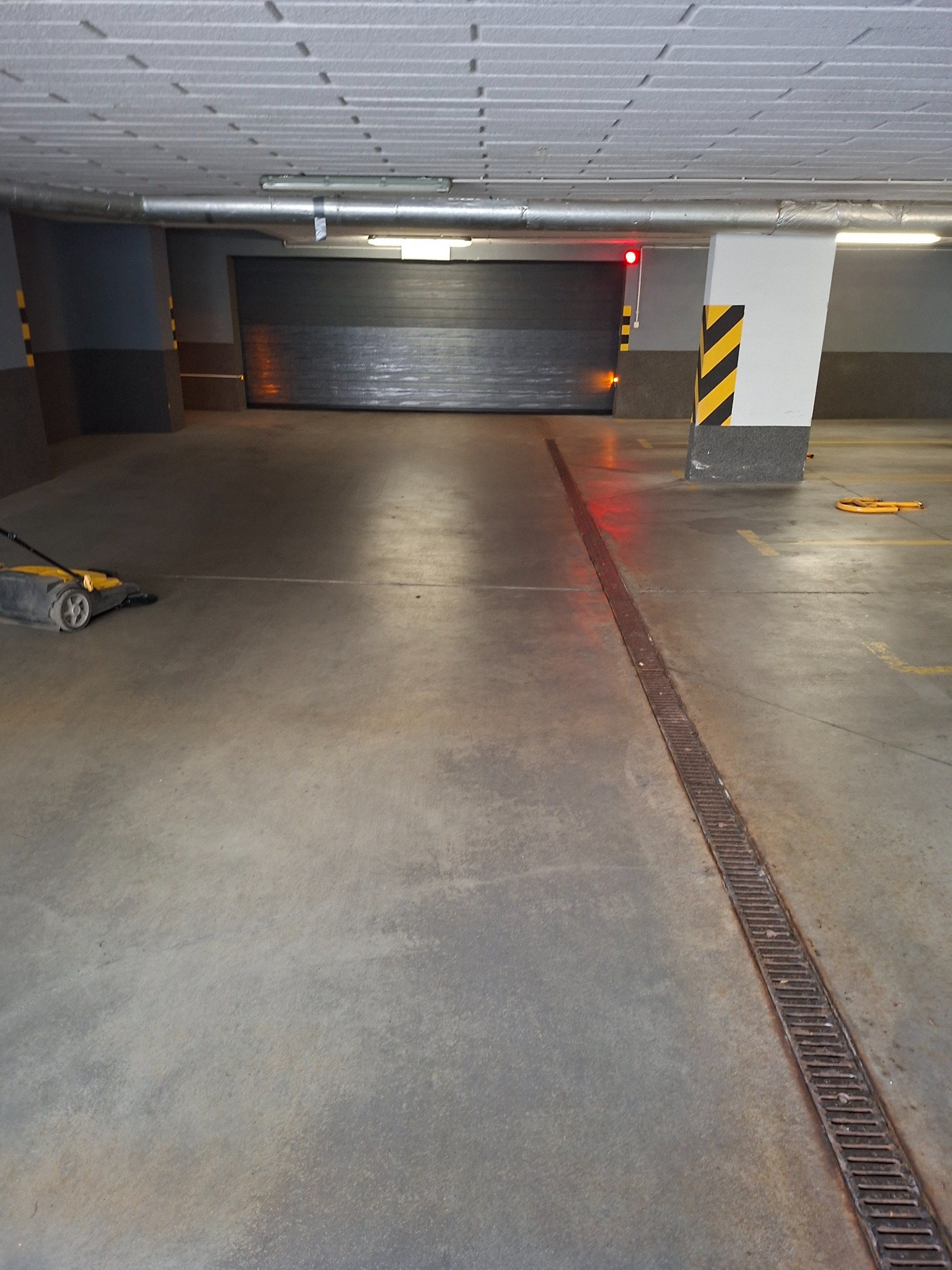 Wnętrze parkingu podziemnego z betonową posadzką i maszyną do czyszczenia podłóg. Widoczna krata odpływowa i brama garażowa z sygnalizacją świetlną.