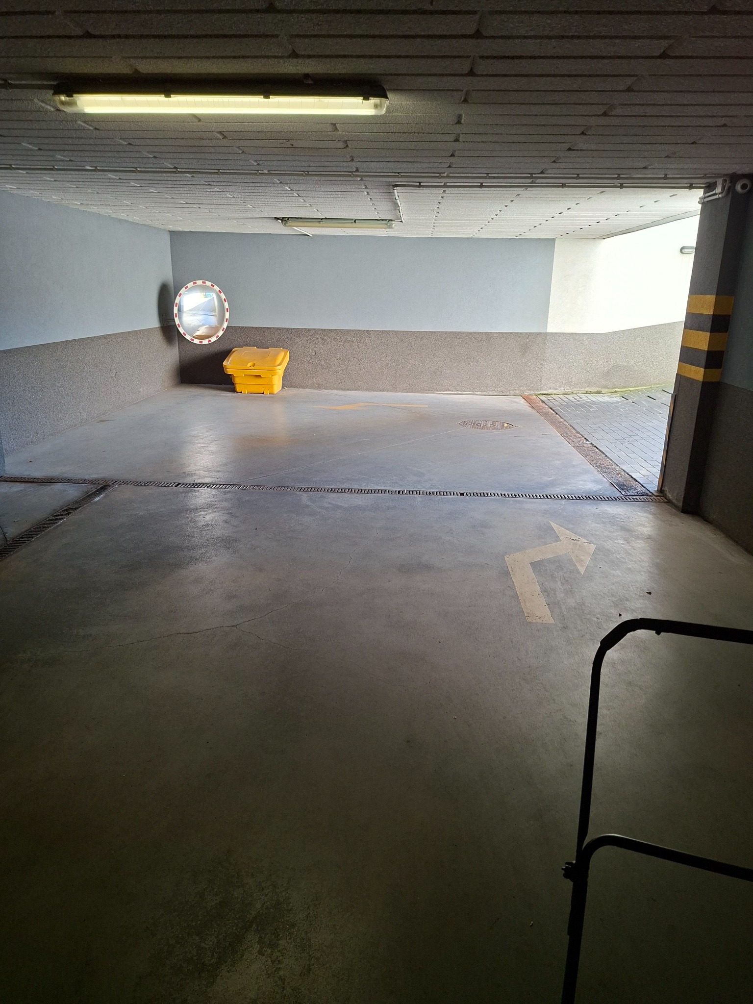 Wnętrze garażu podziemnego z żółtym pojemnikiem na sól drogową, lustrem i strzałką kierunkową na posadzce. Widoczne kratki odpływowe i oświetlenie sufitowe.