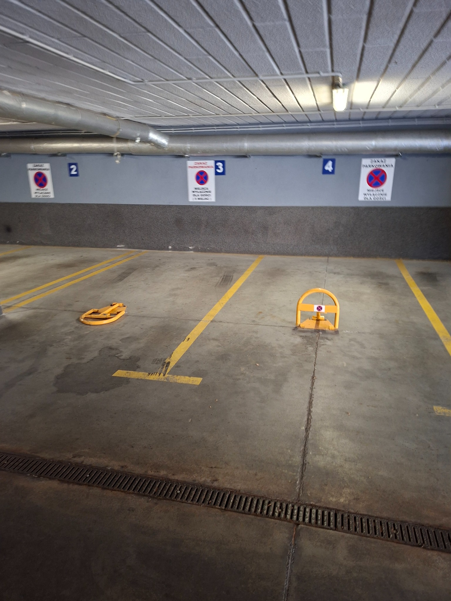 Oznakowane, puste miejsce parkingowe w garażu podziemnym z blokadą. Żółte linie, szara posadzka, znaki zakazu parkowania na ścianie. Widok z góry.