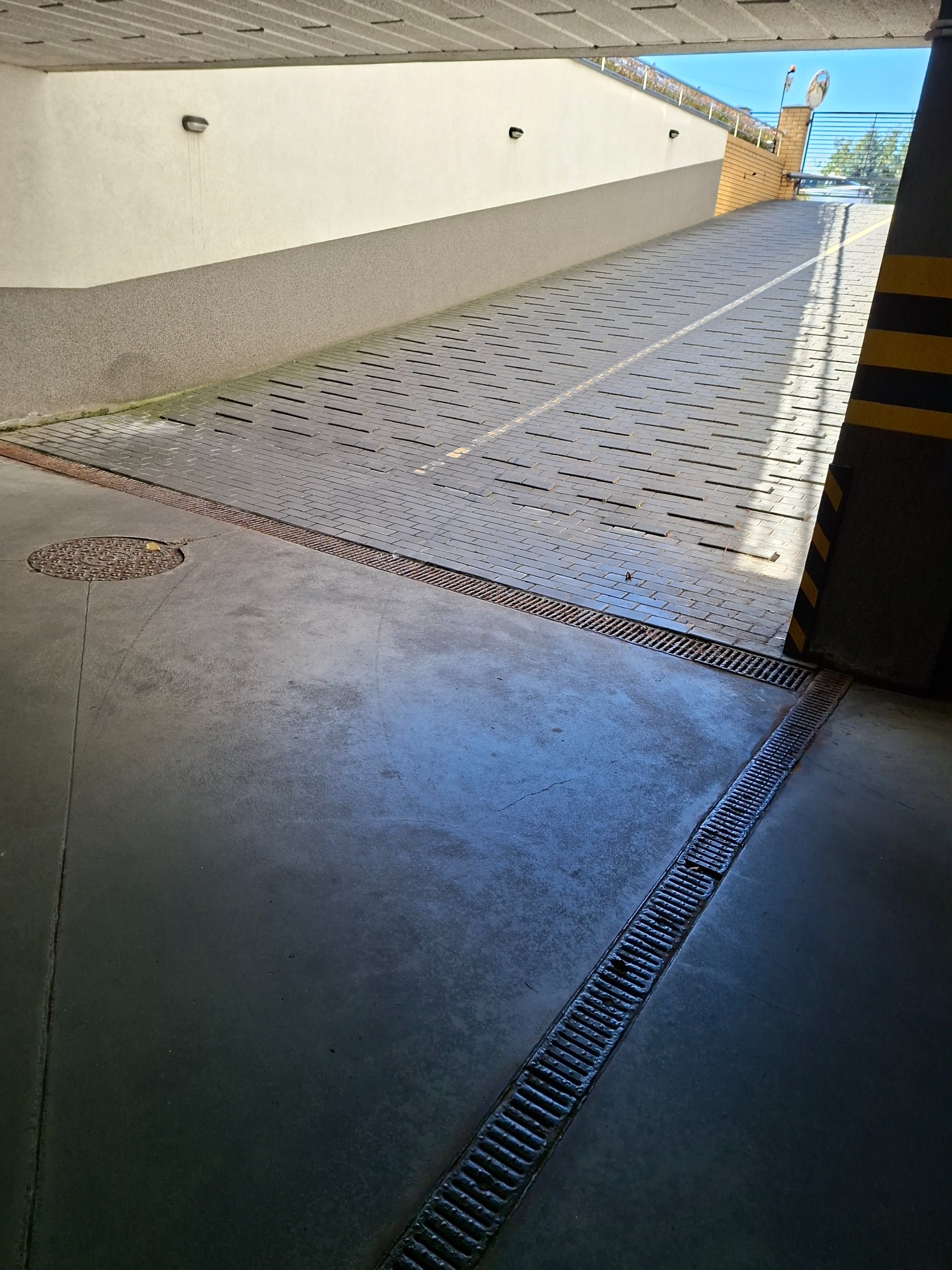 Wjazd do garażu podziemnego z betonową posadzką i kratkami odpływowymi. Widoczny fragment rampy z kostki brukowej i słup z oznaczeniami ostrzegawczymi.