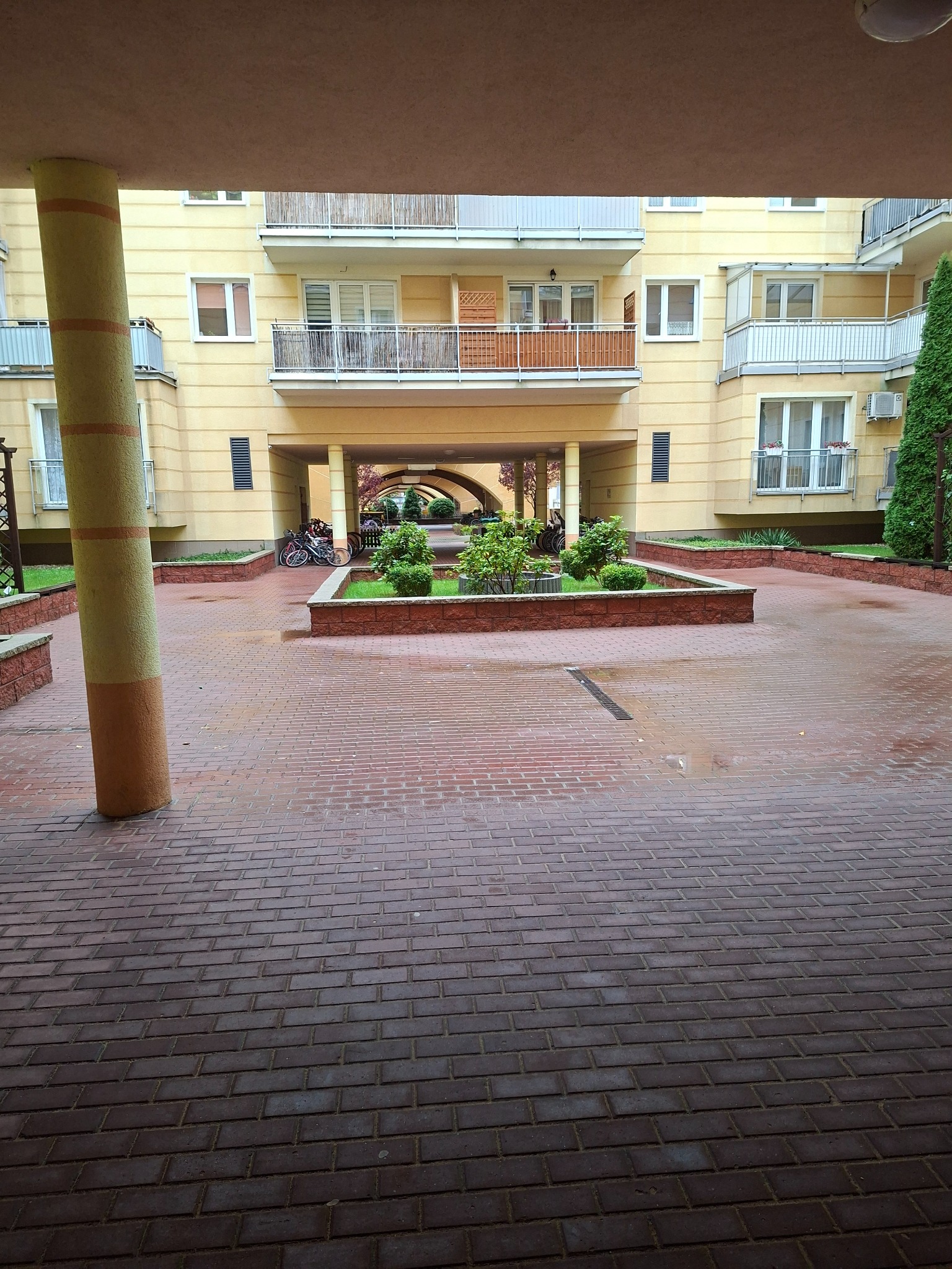 Dziedziniec osiedla z ceglaną nawierzchnią i zielenią w donicach. W tle budynek mieszkalny z balkonami i przejściem łukowym. Widoczny filar i rowery.