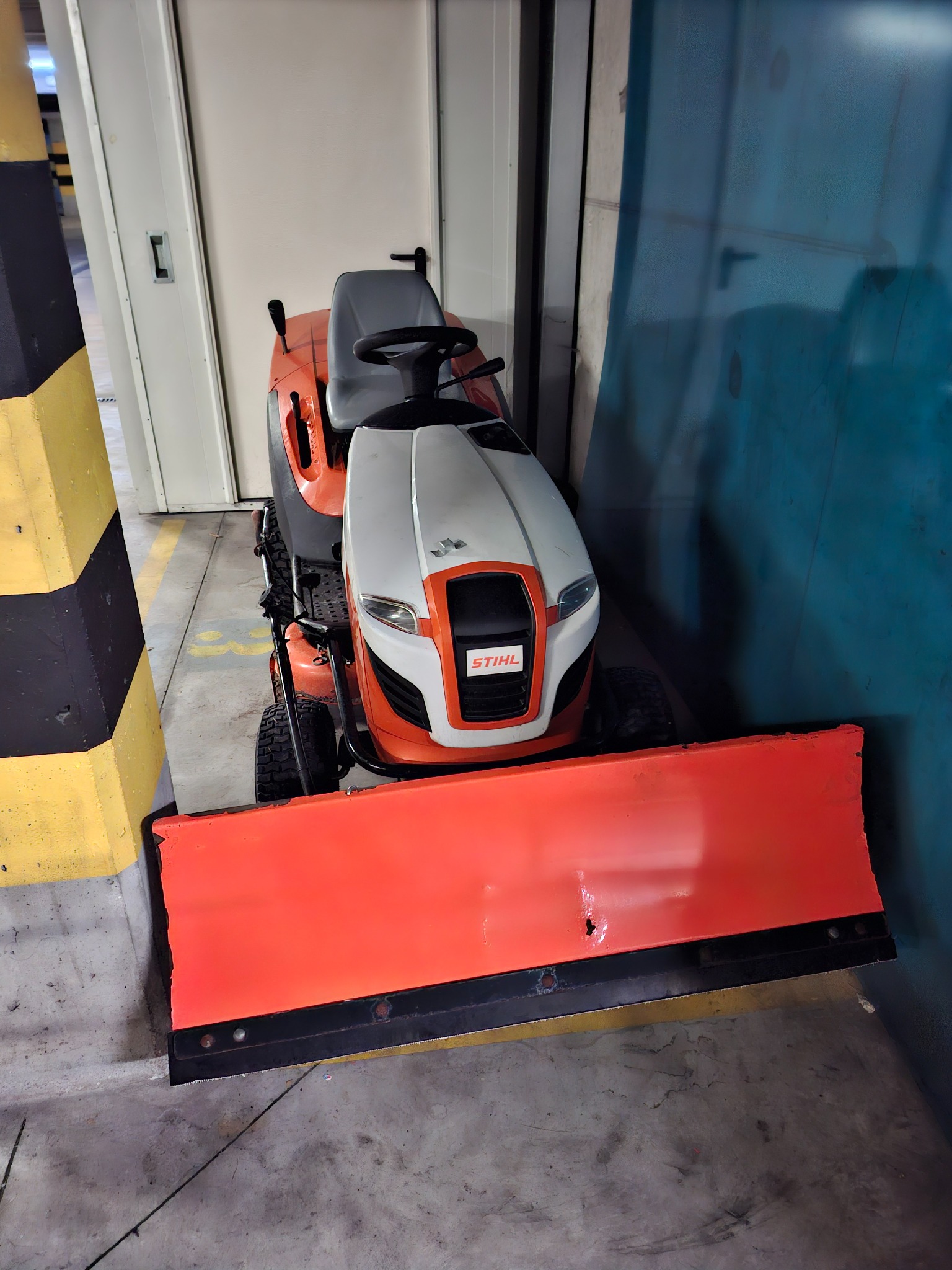 Traktorek Stihl z pomarańczowym pługiem do odśnieżania, zaparkowany w garażu podziemnym, obok słupa z czarno-żółtymi pasami i niebieskiej ściany.