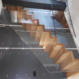 Nowoczesne schody z drewnianymi stopniami, szklaną balustradą i podświetleniem LED wzdłuż ściany.