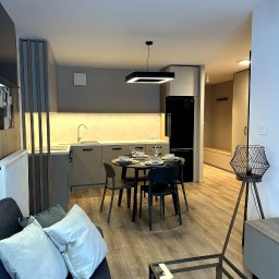 Koral i Kapi Usługi Stolarskie S.C Rafał Koralewski Błażej Błaszczyk - Nowoczesne wnętrze mieszkania z szarą zabudową kuchenną, okrągłym stołem z zastawą i czarną lodówką, widok z salonu.