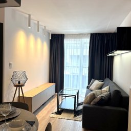 Koral i Kapi Usługi Stolarskie S.C Rafał Koralewski Błażej Błaszczyk - Nowoczesny salon z szarą sofą, stolikiem kawowym ze szklanym blatem, drewnianą szafką RTV i lampą na trójnogu, oświetlony listwą LED na ścianie.