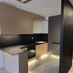 Koral i Kapi Usługi Stolarskie S.C Rafał Koralewski Błażej Błaszczyk - Nowoczesna kuchnia z beżowymi szafkami górnymi i dolnymi, czarnym blatem oraz ścianą nad blatem z efektem kamienia, oświetlona listwą LED pod szafkami, z elementami wykończenia w postaci pionowych...