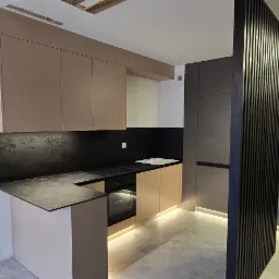 Nowoczesna kuchnia z beżowymi szafkami górnymi i dolnymi, czarnym blatem oraz ścianą nad blatem z efektem kamienia, oświetlona listwą LED pod szafkami, z elementami wykończenia w postaci pionowych...