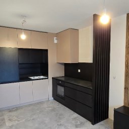 Koral i Kapi Usługi Stolarskie S.C Rafał Koralewski Błażej Błaszczyk - Nowoczesna kuchnia z beżowymi górnymi szafkami i czarnymi dolnymi, drewniana ściana w tle, surowy stan wykończenia podłogi.