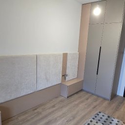 Koral i Kapi Usługi Stolarskie S.C Rafał Koralewski Błażej Błaszczyk - Nowoczesna sypialnia z zabudową: szara szafa na wymiar z długimi, czarnymi uchwytami, tapicerowany panel ścienny z beżowymi akcentami i wbudowanymi gniazdkami, widok na korytarz przez otwarte drzwi...