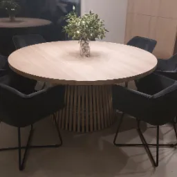 Okrągły stół z jasnego drewna na ażurowej podstawie, otoczony sześcioma ciemnoszarymi krzesłami z metalowymi nogami, na stole wazon z dekoracją roślinną i srebrnymi kulami, w tle fragment aneksu...