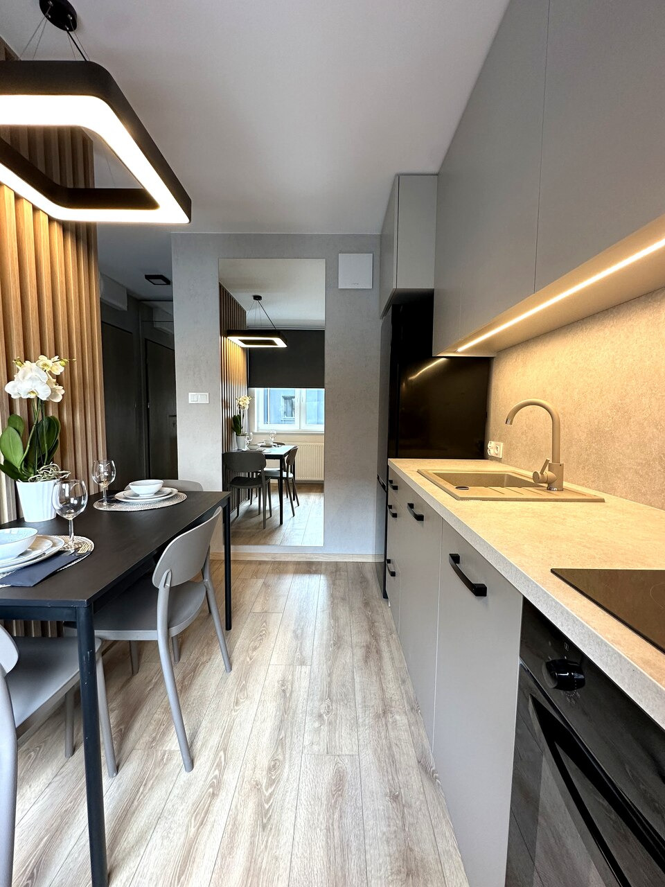 Wnętrze nowoczesnej kuchni z szarymi szafkami, drewnianym blatem, czarnym sprzętem AGD i stołem jadalnym z nakryciem, oświetlone ledowym panelem sufitowym.