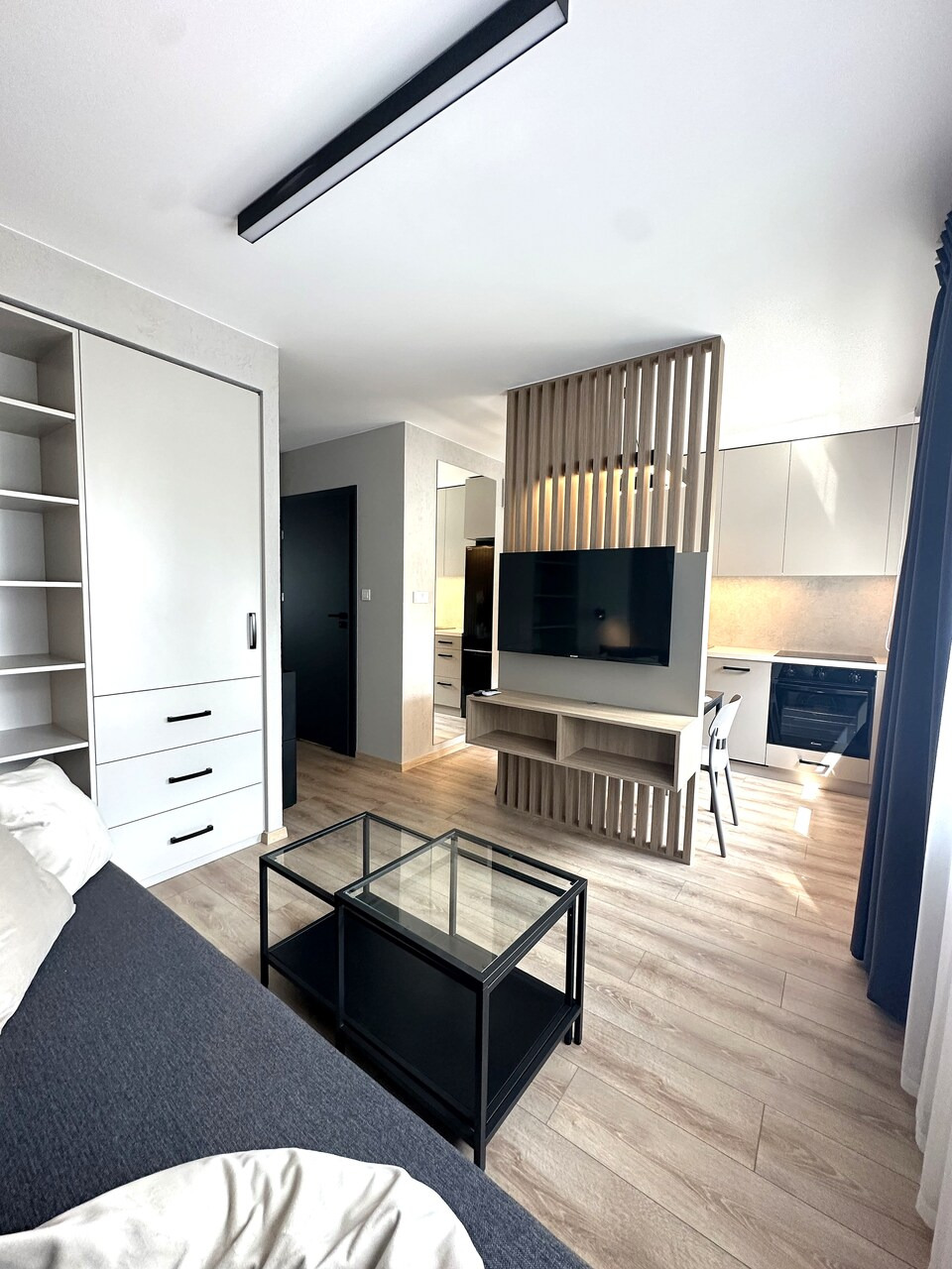 Jasne wnętrze mieszkania z szafą z białymi frontami, szarą sofą, stolikiem kawowym z czarną ramą i szklanym blatem, drewnianą ażurową ścianką działową z telewizorem oraz aneksem kuchennym w tle.