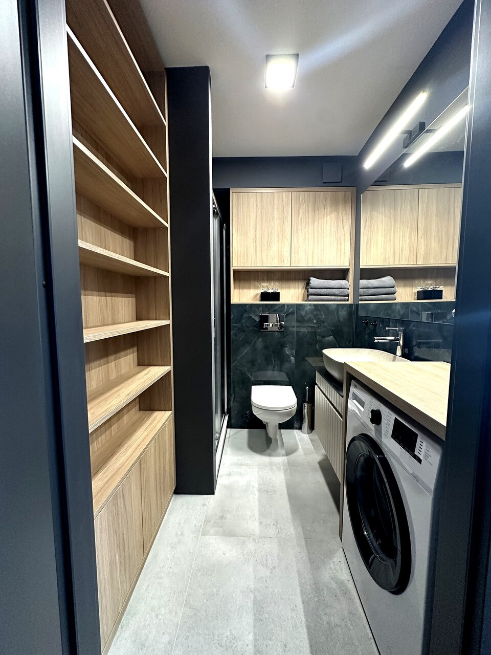 Wnętrze łazienki z zabudową meblową z jasnego drewna, obejmującą otwarte półki i szafki, z pralką pod blatem oraz toaletą w tle, wykończone w ciemnej tonacji.